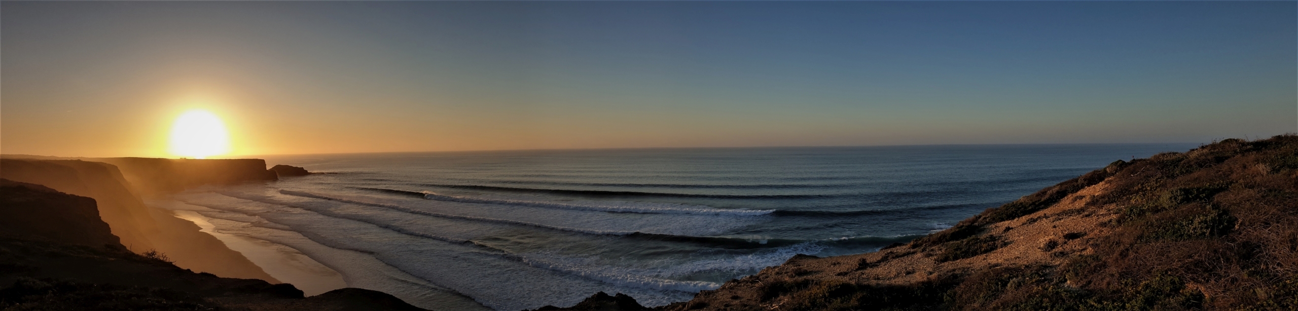 Sonnenuntergang über dem Atlantik in Portugal mit hohen Küstenklippen, Sandstrand und Wellen.