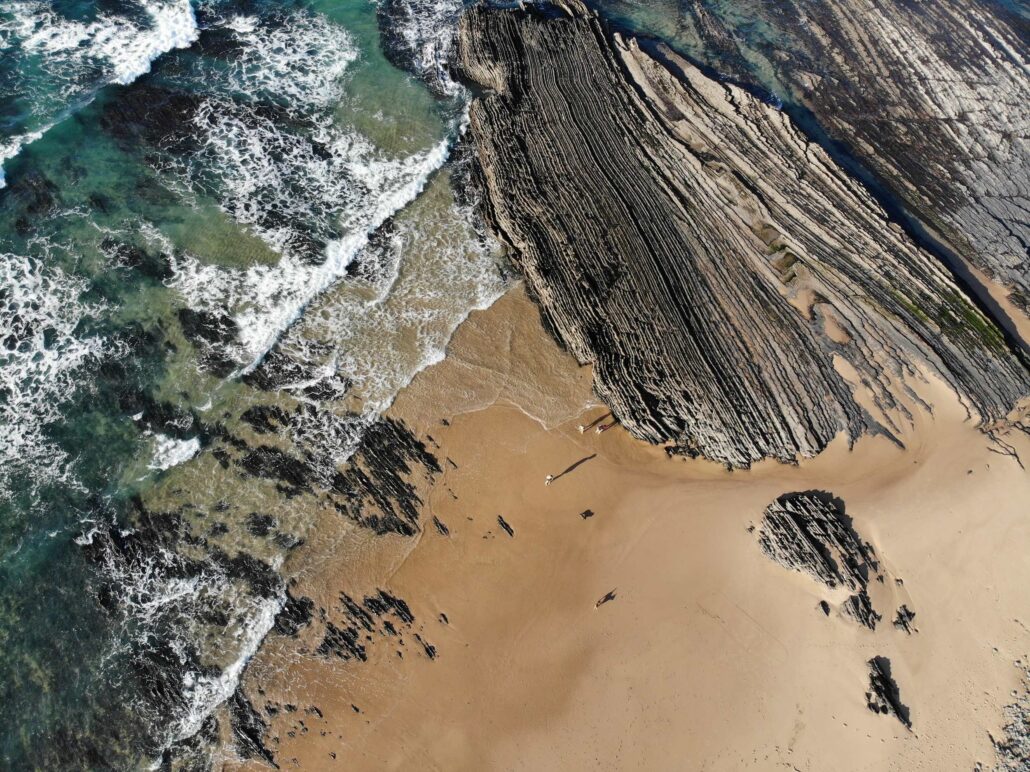 Senkrechte Drohnenaufnahme eines portugiesischen Atlantikstrands mit türkisfarbenem Wasser, Wellen, Sand und markanten Felsformationen.