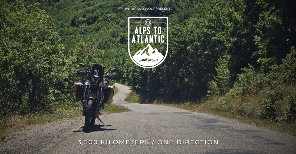 Reisemotorrad auf einer einsamen, von Bäumen gesäumten Straße mit dem Filmlogo A2A – Alps to Atlantic.