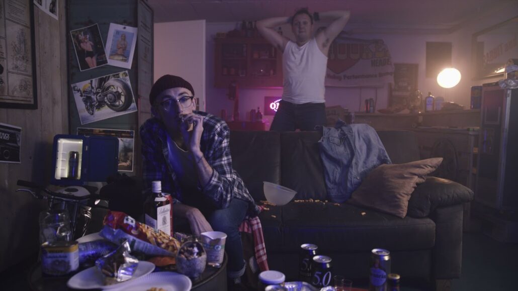 Zwei Schauspieler in einer chaotischen Wohnzimmerkulisse mit Couch, Snacks und Getränken.
